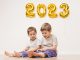 Glückliche Kinder sitzen auf Boden – an der Wand goldene Luftballons 2023