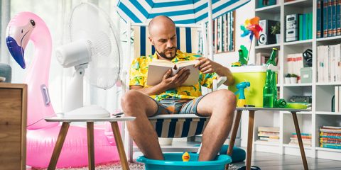 Kühle Wohnung an heißen Tagen – Mann sitzt mit Fußbad und Sonnenschirm im Wohnzimmer