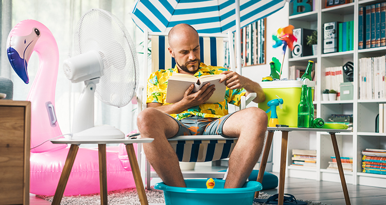 Kühle Wohnung an heißen Tagen – Mann sitzt mit Fußbad und Sonnenschirm im Wohnzimmer